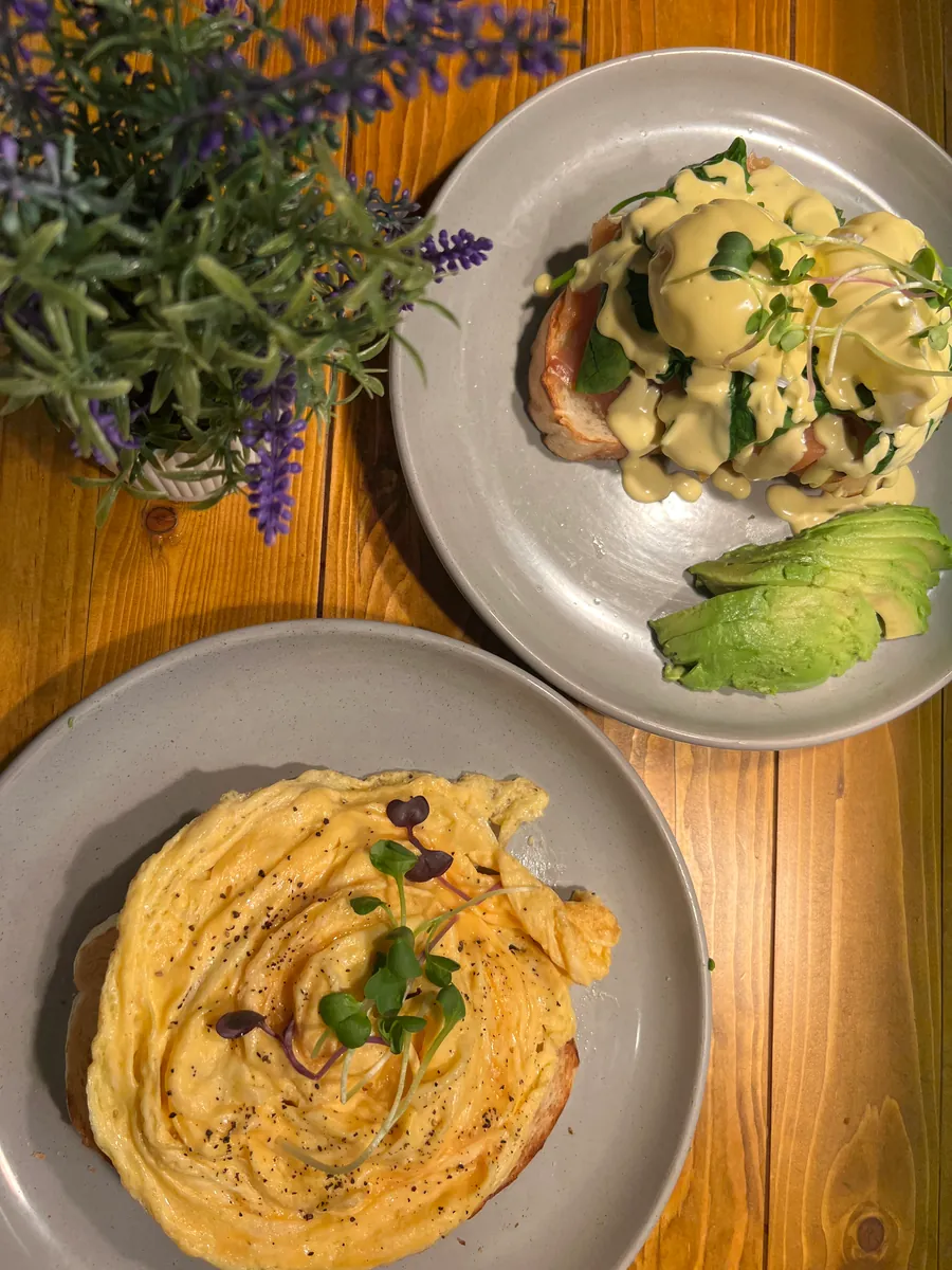 Eggs benny with avocado beside a plate of scrambled eggs on toast