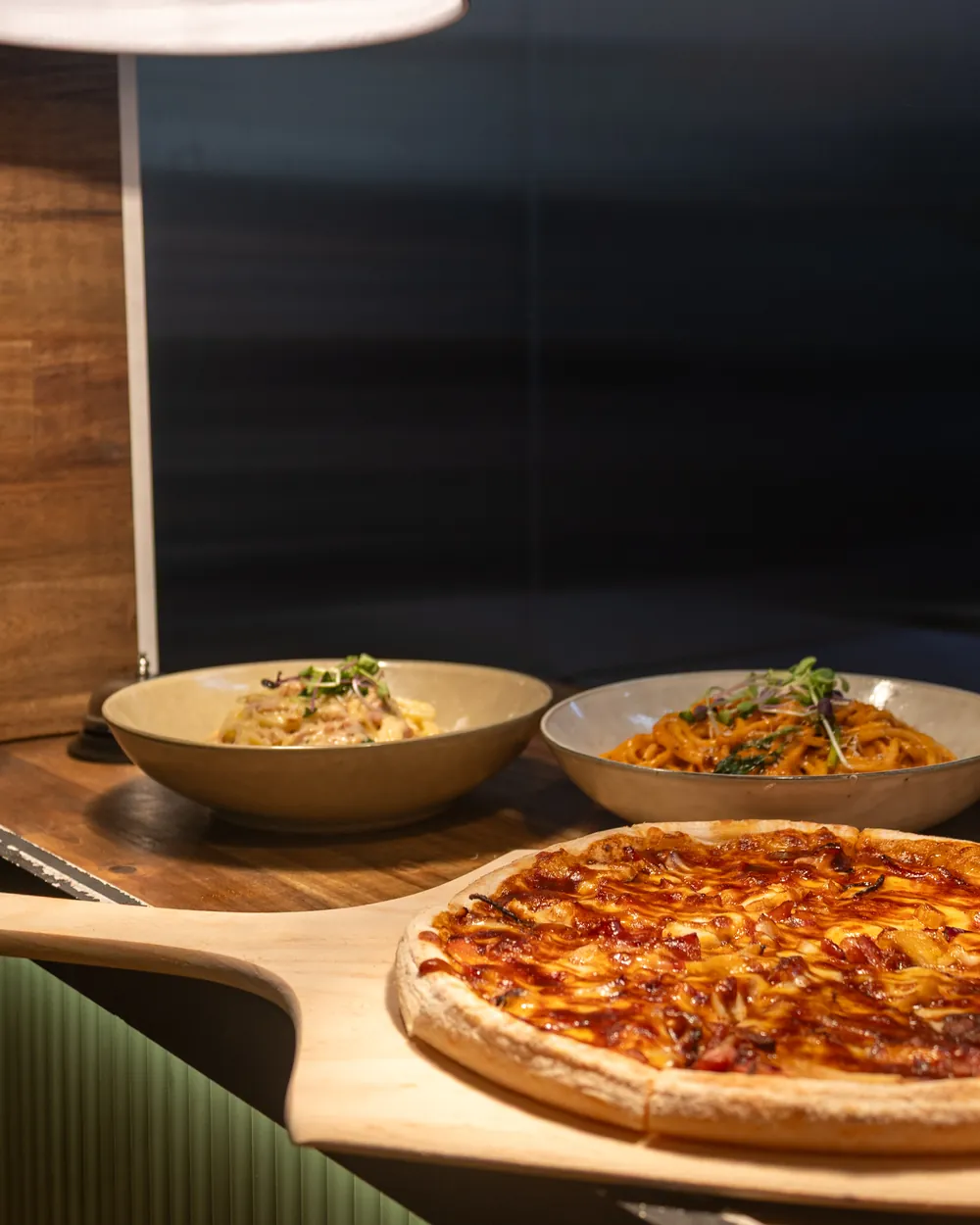 A pizza and two bowls of pasta waiting under the kitchen lamps
